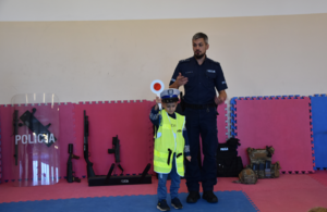Policjant oraz uczeń w kamizelce odblaskowej z napisem Policja oraz tarczą w ręku. Na dalszym planie elementy policyjnego sprzętu