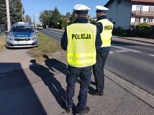 Dwóch policjantów ruchu drogowego w kamizelkach odblaskowych stojących przy drodze. W tle radiowóz.