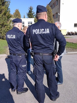 Na tle budynku szkoły dwóch policjantów stojących tyłem. Przed nimi kobieta i mężczyzna.
