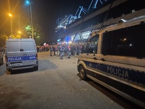 Policyjne radiowozy pod stadionem, w tle widoczni kibice