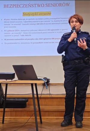 Umundurowana policjantka trzymająca w ręku mikrofon. Obok niej stolik na którym jest komputer, a za nią ekran, na którym jest wyświetlana prezentacja odnośnie bezpieczeństwa seniorów.