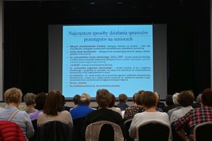 Grupa osób siedzących na krzesłach, osoby są tyłem, przed nimi ekran na którym jest wyświetlana prezentacja odnośnie niebezpieczeństw seniorów