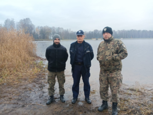 umundurowany funkcjonariusz Policji wraz z dwoma umundurowanymi strażakami Straży Rybackiej na tle stawu