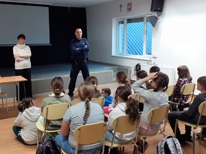 20241112_08105Grupa dzieci siedzących na krzesłach. Przed nimi stoi policjant i kobieta w stroju cywilnym.