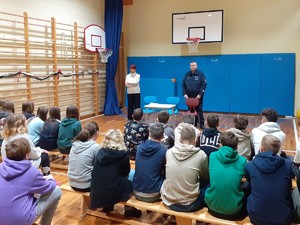 Młodzież, która siedzi na ławkach. Osoby sa tyłem. Przed nimi stoi umundurowany policjant i kobieta w stroju cywilnym.