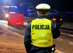 Miejsce śmiertelnego wypadku drogowego  Na zdjęciu widać stojącego tyłem umundurowsnego policjanta  W tle rozłożony parawan wóz strażacki oraz samochód cywilny