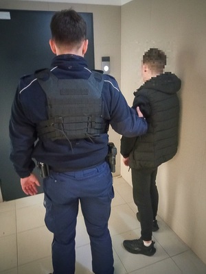 Umundurowany policjant trzymający za rekę zatrzymanego mężczyznę, który jestubrany na czarno