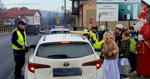 Na zdjęciu po lewej stronie policjant obok samochodu osobowego koloru białego, po prawej grupa dzieci i Mikołaj.