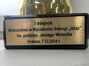 Zbliżenie na tabliczkę znajdująca się na pucharze. wygrawerowany napis "I miejsce Mistrostwa w Wyciskaniu Sztangi "leżąc" im. podinsp. Jerzego Wołocha Kraków, 7.12.2024 r.".