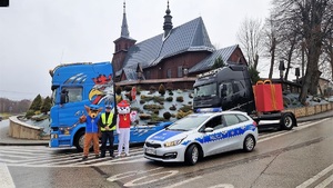 Na tle kościoła dwa samochody ciężarowe, na jednym z nich jest duży czerwony prezent. Obok stoi radiowóz i umundurowany policjant w osobami przebranymi za postacie bajowe, za psy
