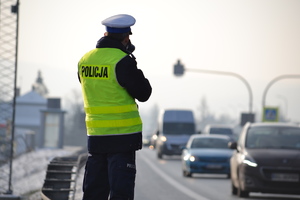 Policjant ruchu drogowego kontroluje ręcznym miernikiem prędkość pojazdów. W tle przejście dla pieszych i przechodząca przez nie kobieta