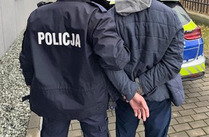 Umundurowany policjant trzymający zatrzymanego mężczyznę. Osoby są tyłem.