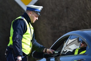 Policjant ruchu drogowego stoi przy samochodzie osobowym i bada siedzącego w środku kierowcę urządzeniem eletronicznym na zawartość alkoholu w wydychanym powietrzu