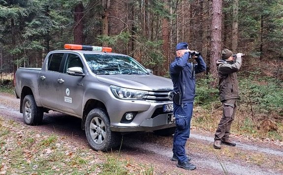 policjant i strażnik leśny patrzą przez lornetki, obok pojazd służbowy Straży Leśnej