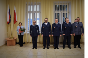 Kadra kierownicza Komendy Powiatowej Policji w Bochni, stojąca z bukietem kwiatów kobieta oraz biało czerwone flagi