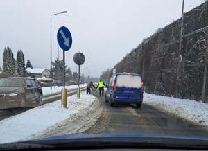 umundurowany plicjant pomaga przeprowadzic osobe poruszającą sie o balkoniku przez jezdnię w tle dwa samochody