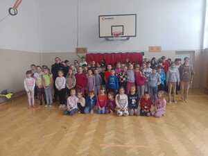 Duża grupa dzieci stoi z razem z policnatem na hali sportowej.