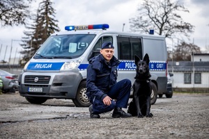 policyjny przzewodnik wraz z psem służbowym w tle radiowóz