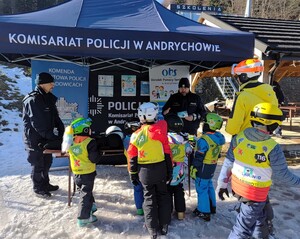 Pod namiotem z napisem Komisariat Policji w Andrychowie stoi dwóch policjantów. Przed nimi grupa dzieci w strojach sportowych zimowych. W tle plakaty i banery.