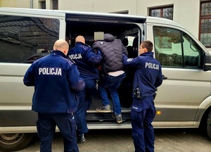 Na zdjęciu trzech umundurowanych policjantów, którzy wprowadzają do samochodu zatrzymanego mężczyznę ubranego w niebieskie dżinsy i czarna kurtkę. W tle budynek.