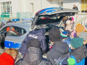 Dzielnicowy schylony przy wnetrzu radiowozu i prezentujący jego wyposarzenie przy słuchających go dzieciach