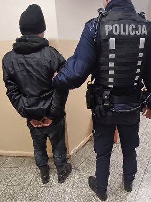 Po prawej stronie umundurowany policjant trzymajacy za rękę zatrzymanego mężczyznę który ma założone kajdanki na ręce trzymane z tyłu  Mężczyzna ubrany jest na