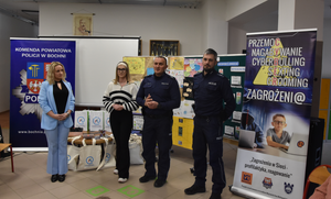 Umundurowani policjanci, dyrekcja oraz praciwnik szkoły. Na zdjęciu widoczne banery Komendy Powiatowej Policji w Bochni oraz akcji Zagrożenia w sieci
