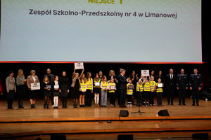 Na scenie laureaci konkursu odblaskowa szkoła z nagrodami i dyplomami wraz ze stojącymi policjantami oraz MWicekuratorem Oświaty