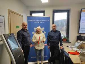 Dwóch policjantów stoi razem z seniorką w pomieszczeniu ZUS na tle policyjnego baneru.