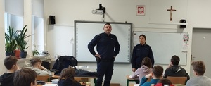 Policjant i Policjantka stojący w klasie szkolnej przed kilkoma uczniami