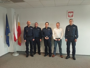 Czterech policjantów w mundurze oraz mężczyzna w białej koszuli stajacy na tle białej ściany z obrazem godła Polskiego, po lewej stronie 3 flagi.