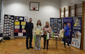 Uczennica z dyplomem za udział w konkursie. Umunudorwany policjant oraz dyrekcja szkoły. Na zdjęciu widoczne są tablice z pracami konkursowymi oraz banery.