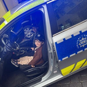 Dwóch małych chłopców siedzacych z przodu w radiowozie. Na głowie mają policyjne czapki. Drzwi od strony loerowcy sa otwarte.