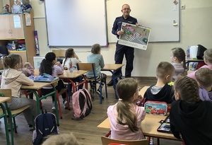 1 policjant w sali lekcyjnej, przy pomocy tablicy edukacyjnej, wyjaśnia dzieciom jak prawidłowo przechodzić przez jezdnię