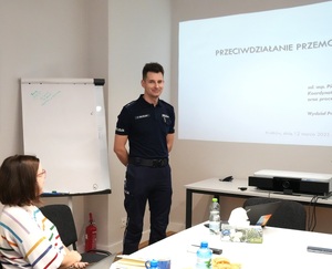 2 Policjant prowadzi szkolenie w tle widać prezetację przy stole jedna z uczestniczek szkolenia