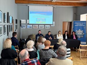 Pred stołem siedzą dwaj policjanci oraz trzy kobiety w stroju cywilnym. Przed nimi grup osób siedzacych na krzesłach. W tle ekrn na ktrórym jest wyświetlana prezentacja. Po prawej stronie policyjny baner2mp
