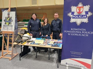 Dwoje policjantów oraz pracownik cywilny stoi przy stoisku Policji na targach pracy.