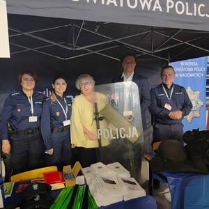 Na zdjęciu po lewej diwe policjantki, dalej kobieta i mężczyzna w stroju cywilnym przed którymi jest policyjna tarcza, po lewej policjant. Przed nimi na stoliku różne gażdżety.