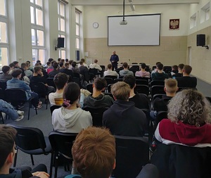 Na zdjęciu grupa uczniów siedzących na krzesłach. Osoby sa tyłem. Przed nimi umundurowany policjant.