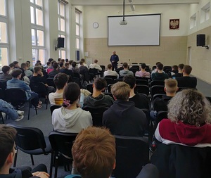 Na zdjęciu grupa uczniów siedzących na krzesłach. Osoby sa tyłem. Przed nimi umundurowany policjant.