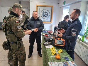 Po lewej stronie dwóch chłopaków w mundurach wojskowych. Obok nich dwaj umundurowani policjanci,jeden stoi za stolikeim na którym sa różne gażdżety.