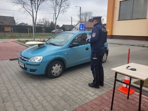Niebieski samochód osobowy, na którym na masce przymocowany jest talerz na którym jest piłeczka. Obok policjant.