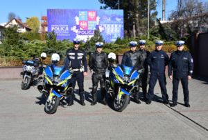 Policjanci obok oznakowanych motocykli. a nimi widoczna jest tablica z logo jednostki