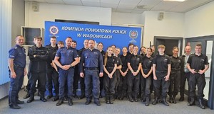 Na zdjęciu uczniowie klasy policyjnej w czarnych mundurach, trzech umundurowanych policjantów. W tle baner z napisem Komenda Powiatowa Policji w Wadowicach.