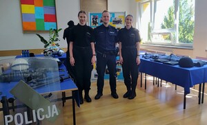 Umundurowany policjant, po prawej i po lewej uczennice w mundurach szkolnych. W tle przygotowana gazetka, na której są zdjęcia. Na stolikach widać również poukladany sprzęt policyjny.