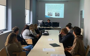 Po prawej i lewej stronie, przy sole, siedzą młode osoby. Przed nimi policjantka, a za nią na ekranie wyświetlana prezentacja.