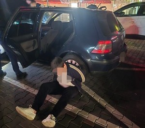 Zatrzymany mężczyzna siedzący na ziemi z kajdankami oparty o niebeski pojazd