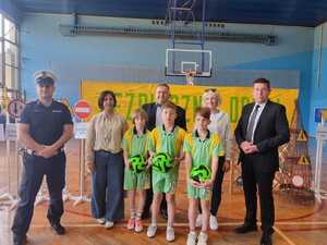 Od lewej stoi umundurowany policjant, dwie kobiety i dwóch mężczyzn w strojach cywilnych, a przed nimi trzech chłopców w zielonych strojach sportowych, któtzy trzymają w rękach piłki.