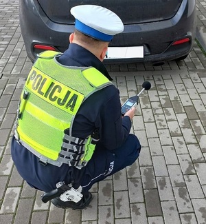 Umundurowany policjant ruchu drogowego w odblaskowej kamizelce z napisem Policja za pomocą sonometru sprawdza  normy hałasy emitowane przez pojazd.