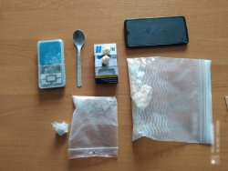 dwie foliowe torebki z białą substancją, waga elektroniczna, zbrylona substancja na opakowaniu po papierosach, łyżeczka oraz telefon komórkowy